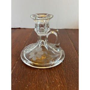 Peut inclure: Bougeoir en verre transparent avec une poignée et un motif floral gravé. Le bougeoir a une base ronde et un dessus détaillé pour tenir une bougie. La poignée est une petite boucle. Le motif gravé représente une marguerite.