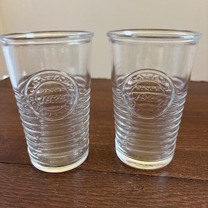 May include: Two clear glass tumblers with a ridged design and a circular emblem that reads "SINCE 1825 QUALITY". The glasses are cylindrical with a slightly flared rim. The glasses are on a brown surface.