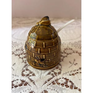 May include: A brown ceramic honey pot shaped like a beehive, with a matching lid and a white spoon. The pot features embossed details and bee illustrations. The honey pot is a kitchenware item.