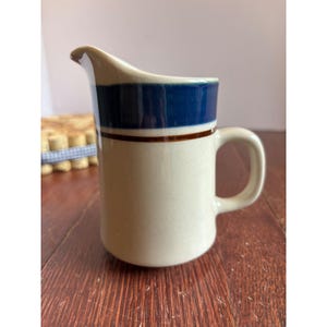 May include: A cream-coloured ceramic jug with a dark blue band at the top and a brown stripe below it. The jug has a curved spout and a rounded handle. The jug is on a wooden surface.