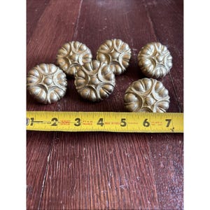Op de afbeelding: Zes antieke messing kastknoppen met een bloemmotief liggen op een houten oppervlak. Een gele meetlint ligt in de buurt van de knoppen. De knoppen hebben een diameter van ongeveer 5 cm.