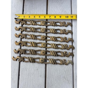 May include: Six identical antique brass-toned metal furniture pulls with an elongated, ornate scrollwork design. Each piece features a central mounting hole and measures approximately 11 inches in length as shown by the yellow measuring tape.