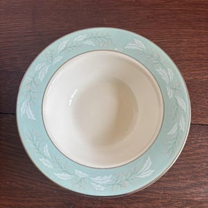May include: A round, off-white ceramic bowl with a light teal border. The border features a repeating pattern of white leaves and small pink dots. A thin gold band accents the inner and outer edges of the teal border. The bowl sits on a dark wood surface.