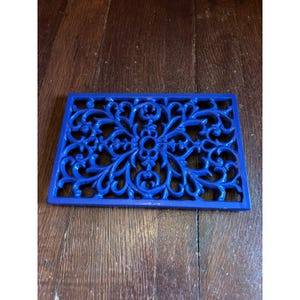 May include: A rectangular cast iron trivet featuring a vibrant blue finish and an intricate, symmetrical openwork scroll pattern. The design consists of interconnected floral-inspired curves and loops, creating a decorative lattice structure.