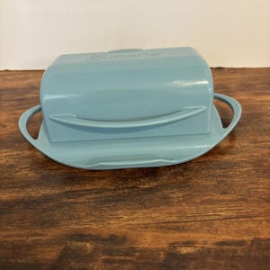Op de afbeelding: Een lichtblauwe botervloot met een deksel en handvatten. De schaal heeft een rechthoekige vorm met afgeronde randen en een bijpassende basis. Het woord "Butterier" staat op het deksel.