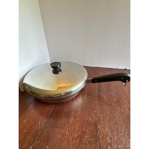 May include: A round stainless steel skillet with a polished finish and a matching lid. The pan features a long, black heat-resistant handle with a small metal loop at the end for hanging, and the lid has a central black knob for easy lifting.