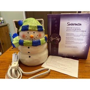 Peut inclure: Un chauffe-parfum Scentsy en forme de bonhomme de neige en céramique avec un bonnet et une écharpe en tricot vert et bleu. Le bonhomme de neige a un nez en carotte et des yeux en bouton noirs. Un cordon d'alimentation blanc et la boîte du produit sont visibles.