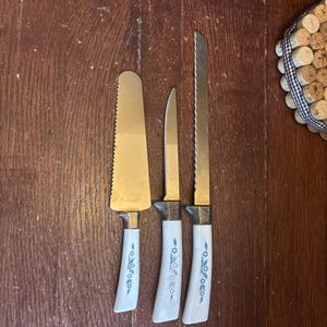May include: A set of three vintage serving knives with gold blades and white handles. The handles are decorated with a blue floral pattern. The knives are arranged on a dark wooden surface.