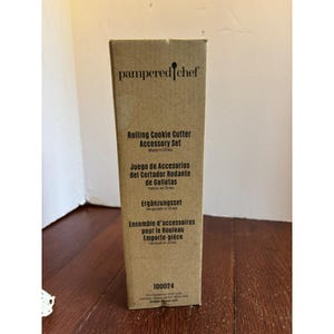 以下が含まれることがあります： Pampered Chef ローリングクッキーカッターアクセサリーセット用の茶色の段ボール箱。箱には英語、スペイン語、ドイツ語、フランス語のテキストがあります。箱には番号100024も印刷されています。