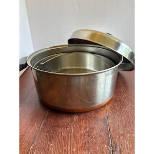 May include: A stainless steel cooking pot with a copper-colored base and a matching lid. The pot features a removable internal steamer insert with a perforated bottom and black side handles. The exterior surface shows light surface scratches.