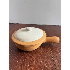 May include: A small, round, terracotta-coloured ceramic cooking pot with a single handle extending from the side. The pot features a smooth, off-white glazed lid with a small, centred knob handle, contrasting with the unglazed, matte exterior.