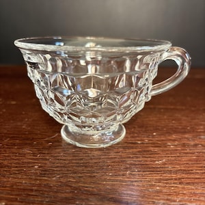 May include: Clear glass teacup with a diamond pattern. The teacup has a handle and sits on a small, round base. The glass is transparent, allowing light to pass through. The teacup is designed for serving hot beverages.