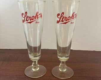 May include: Two clear, stemmed beer glasses with the red "Stroh's Beer" logo. The glasses have a slender, flared shape and a round base. The logo is in a cursive font with a red underline.