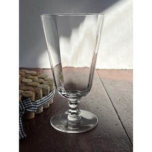 May include: A clear glass goblet with a faceted, tapered bowl and a multi-tiered, decorative stem resting on a circular base. The glass features vertical panels that create a geometric silhouette, reflecting light through its transparent surface.