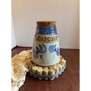May include: A ceramic popcorn jar with a blue rim and floral design, topped with a cork stopper. The word "POPCORN" is displayed on the front. It sits on a coaster made of corks and a blue and white checkered ribbon.