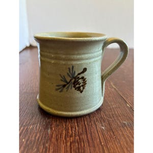 May include: A light tan ceramic mug with a smooth, speckled finish and a single handle. The front of the mug features a hand-painted brown pinecone and blue pine needles. The mug has a slightly flared rim and a subtle horizontal ridge detail.