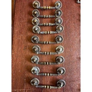 May include: A collection of eight antique-style brass drawer pulls arranged in a vertical column. Each pull features a central horizontal bar with ribbed detailing and two circular mounting plates at the ends, decorated with a sunburst pattern.