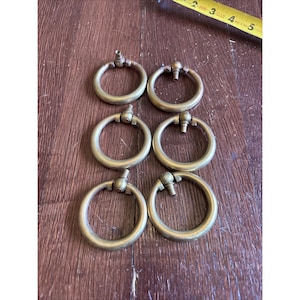 May include: Six antique brass ring pulls for furniture. Each pull has a circular ring attached to a decorative post. The pulls are arranged on a wooden surface. The pulls are a warm, golden color.