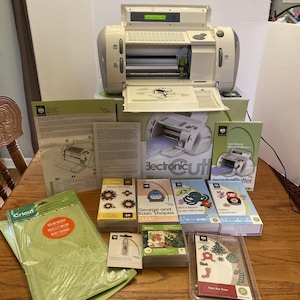 Puede incluir: Una máquina de corte electrónica Cricut con varios accesorios, incluyendo tapetes de corte, cartuchos y manuales. La máquina es blanca y gris, con un panel frontal abierto. Los accesorios incluyen cajas con diseños de flores, formas y festivos.
