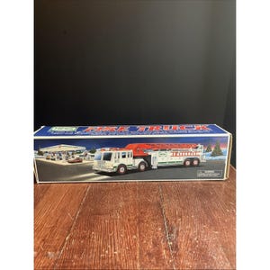 May include: A vintage Hess Fire Truck toy in its original box. The box features a colorful illustration of the fire truck with its ladder extended, a gas station, and a winter scene. The truck is white with red accents and a long ladder.