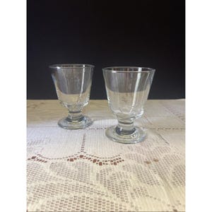 May include: Two clear, stemmed glasses with a flared bowl and a round base. The glasses are made of transparent glass and sit on a white lace tablecloth. The background is a solid black.