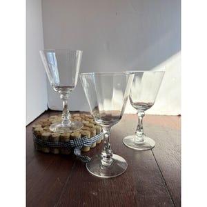 May include: Three clear glass goblets with smooth, wide bowls and faceted, decorative stems. Each glass features a circular base and a transparent finish. The glasses are arranged together, with one resting on a circular cluster of corks.