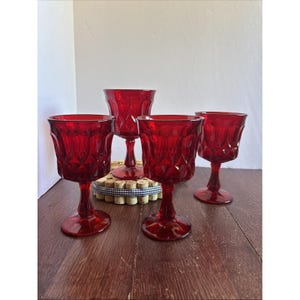 May include: Four ruby red wine glasses with diamond-cut pattern. The glasses have a stemmed base and a wide bowl. The glasses are arranged on a wooden surface, with a white background.