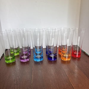May include: A collection of eleven tall, clear drinking glasses arranged in two rows. Each glass features a thick, weighted base tinted in a vibrant color, including shades of lime green, purple, blue, dark blue, orange, and bright red.