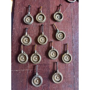 May include: A collection of eleven circular brass drawer pulls, each featuring a central sunburst pattern and a hinged ring handle. Each pull includes a threaded metal bolt extending from the top, designed for attachment to furniture surfaces.