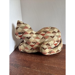 May include: A cat-shaped pillow with a patchwork design in shades of brown, tan, and red. The pillow features a button eye and a curved tail. The fabric is patterned with triangles and small squares.