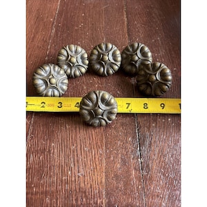 May include: Six antique brass cabinet knobs with a floral design are arranged on a wooden surface. A yellow measuring tape is placed near the knobs. The knobs are approximately 1.5 inches in diameter.