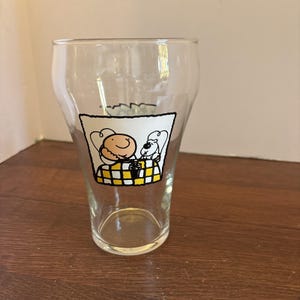 May include: A clear glass pint glass with a cartoon graphic. The graphic features two characters, one with a smiling face and the other resembling a dog, seated on a yellow and black checkered surface. The glass is empty and sits on a wooden surface.