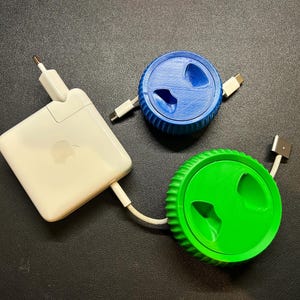 Pode incluir: Um adaptador de energia Apple branco, conectado a um enrolador de cabo azul e verde. Os enroladores de cabo têm um design circular com uma borda canelada e uma ranhura para o cabo. Para carregar dispositivos.