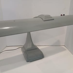 May include: A gray desk lamp with a long, curved shade and a rectangular base. The lamp has a streamlined design with a small decorative element on top of the shade. The lamp is made of metal and has a power cord.