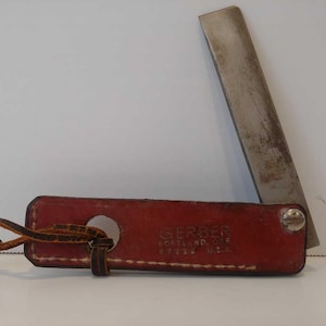 May include: A vintage Gerber knife with a red leather sheath and a metal blade. The sheath has a hole for a leather strap and the text "GERBER PORTLAND, ORE 97223 U.S.A."