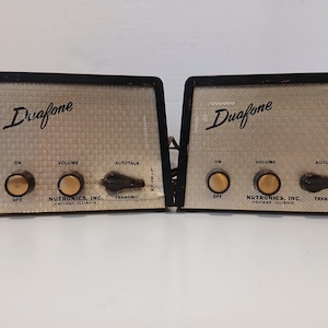 May include: Two vintage Duafone intercom units. Each unit has a black frame, a beige mesh front, and gold-colored knobs for volume and power. The text "Duafone" is displayed prominently on the front.