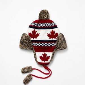 May include: A knit trapper hat featuring a red and white pattern with red maple leaf motifs, dark blue geometric bands, and brown faux fur trim on the ear flaps and pom-poms. The hat includes two red braided ties with faux fur pom-poms.