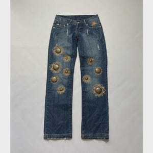 May include: Blue denim jeans featuring a distressed wash and a series of circular, metallic, gold-toned studs arranged in floral-like patterns across the front of the legs. The jeans include a standard button fly, belt loops, and five pockets.