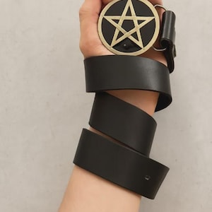 May include: A black leather belt featuring a circular buckle with a gold-toned pentagram design set against a black background. The belt strap is wrapped around the wrist, showcasing the smooth texture of the leather and the metallic buckle.