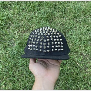 Può includere: Un cappellino da baseball nero caratterizzato da una fitta disposizione di punte metalliche color argento che coprono l'intero pannello frontale e superiore. Il cappellino presenta una visiera piatta e una corona strutturata e uniforme.