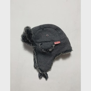 Può includere: Un cappello da trapper in maglia grigio scuro con fodera in pelliccia sintetica e paraorecchie. Il cappello presenta un piccolo dettaglio metallico circolare sul lato e un'etichetta rettangolare rossa con la scritta bianca Kickers.