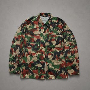 Op de afbeelding: Een overhemd met lange mouwen en knoopsluiting, voorzien van een camouflagepatroon in tinten groen, bruin, zwart en beige. Het overhemd heeft een standaard kraag, twee borstzakken met knoopsluiting en epauletten voor een gestructureerde uitstraling.
