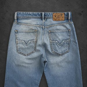 May include: Light blue denim jeans featuring a faded wash, two back pockets with V-shaped stitching, and a tan leather patch on the waistband with the text Diesel Industry. The jeans have a classic five-pocket construction and belt loops.
