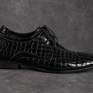 Puede incluir: Un zapato negro con cordones, acabado brillante y textura de piel de cocodrilo. El zapato presenta un tacón bajo, una punta redondeada y cordones negros finos que pasan a través de ojales en la parte superior, mostrando un aspecto pulido.