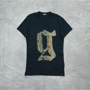 May include: A black short-sleeve V-neck t-shirt featuring a large, stylised letter G graphic on the front. The letter is filled with a textured, multi-coloured snakeskin pattern in shades of green, tan, and brown, with a dark blue interior accent.
