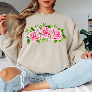May include: A beige sweatshirt featuring a watercolor floral design. The design includes three large pink roses, smaller pink flowers, and green leaves. The sweatshirt is a neutral color and has a crew neck.