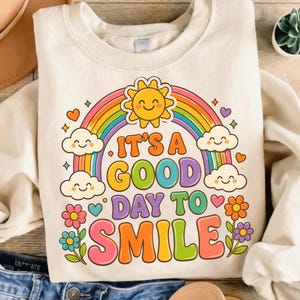 May include: A cream-colored crewneck sweatshirt featuring a colorful graphic print. The design includes a smiling sun, a rainbow, clouds, flowers, and hearts. The text reads, "IT'S A GOOD DAY TO SMILE" in large, multi-colored, rounded bubble letters.