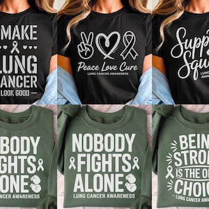 Puede incluir: Una colección de camisetas negras y verde oliva con texto blanco y cintas de concienciación sobre el cáncer de pulmón. Las camisetas muestran frases como "I Make Lung Cancer Look Good", "Peace Love Cure" y "Nobody Fights Alone".
