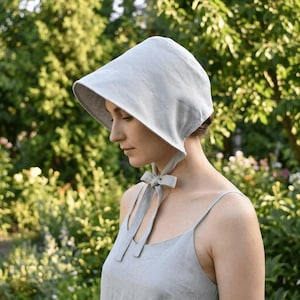 May include: A light gray bonnet made of fabric with a wide, structured brim that frames the face. The headwear features long, matching fabric ties that are knotted securely under the chin, providing a secure fit and a classic, simple silhouette.