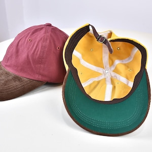Peut inclure: Une casquette de baseball marron avec une visière en velours côtelé marron à côté d'une casquette jaune avec un dessous vert, un bandeau marron, des bandes de couture intérieures blanches et une sangle réglable en cuir marron avec boucle.